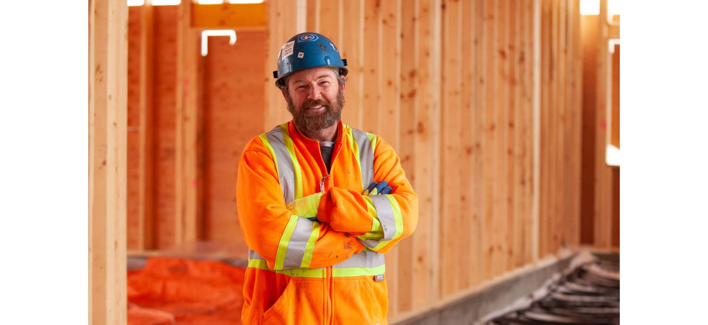 Brad standing with his arms folded wearing high visibility gear and a helmet on a construction site.